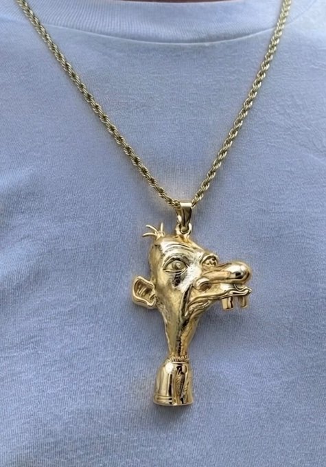 A gold pendant featuring a stylized, cartoonish face with an exaggerated grin, prominent nose, and protruding teeth hangs from a twisted gold chain against a light blue shirt. The pendant's ostentatious, gaudy design serves as a visual "sign" of newfound wealth, complementing the post's playful warning about flaunting profits from $Neegy without spelling it out.