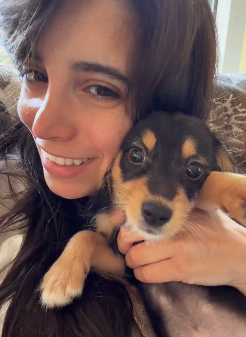 A woman with long dark hair smiles warmly while cradling an adorable black and tan puppy close to her cheek, capturing a sweet "good morning" moment with her furry companion.