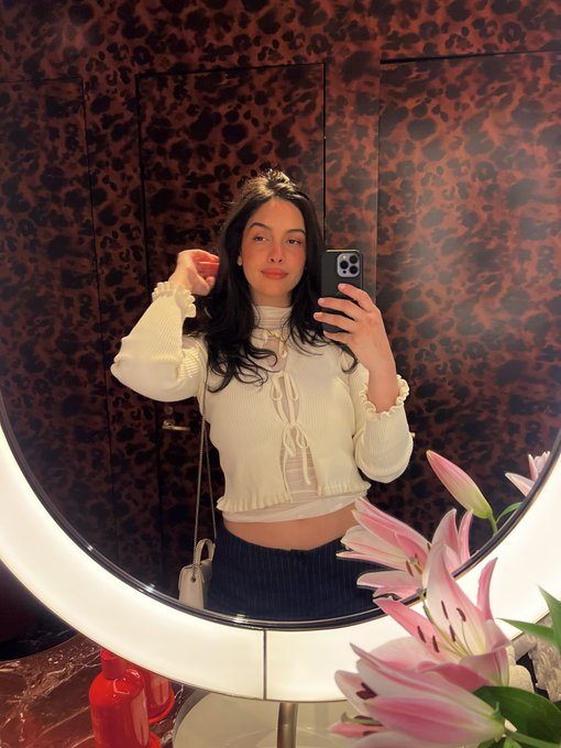 A mirror selfie of a woman with long dark hair wearing a cream-colored ruffled top, holding a smartphone, with pink lilies visible in the foreground.