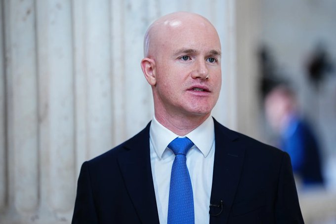 A bald man in a dark suit with a blue tie speaks during what appears to be an interview, with stone columns visible behind him.