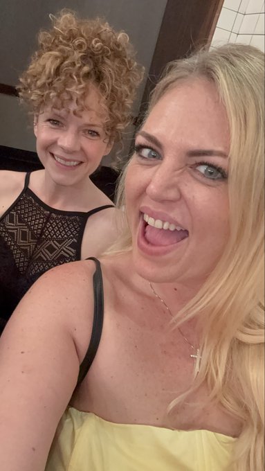 A selfie of two women, one with curly blonde hair in a black lace top and the other with long blonde hair in a yellow top, both smiling widely.