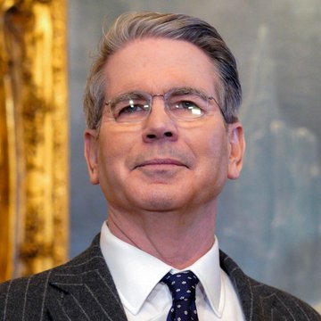 I can see a man in the image with gray hair, wearing glasses, a white dress shirt, and a pinstripe suit with a blue patterned tie. He appears to be looking upward with a slight smile, set against what looks like an ornate interior with gilded architectural details.

Based on the accompanying post, this appears to be Treasury Secretary Scott Bessent, pictured as the article discusses his views that the Federal Reserve should remain open-minded about interest rates, citing AI productivity gains and deregulation as potential tools to manage U.S. inflation.