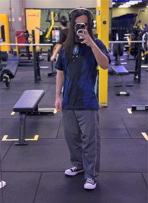 A mirror selfie taken in a gym showing a person wearing a blue and black patterned t-shirt, grey pants, black sneakers, and over-ear headphones, holding a phone to take the photo.
