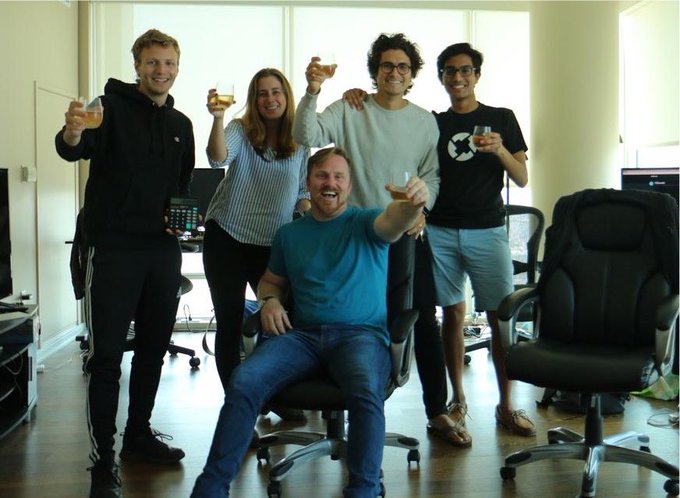 A group of five people pose in an office, raising glasses in a celebratory toast, with one man seated in a chair at the center and four colleagues standing behind him holding drinks. The image complements the post's reflection on Polychain's 10-year journey, capturing the camaraderie of the team that helped turn the once "impossible dream" into reality.