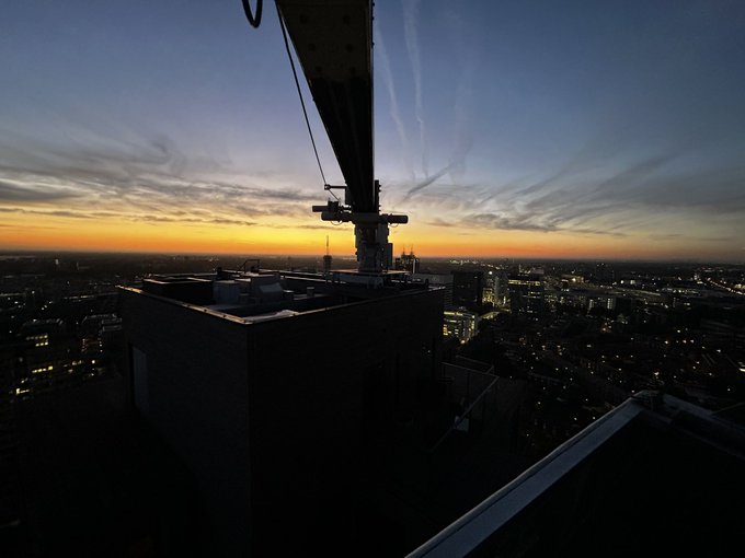 A construction crane silhouetted against a vibrant sunrise over a sprawling cityscape, with building lights still twinkling in the pre-dawn glow. The image reinforces the post's message to "touch some grass," capturing the kind of breathtaking outdoor view and fresh morning atmosphere XRP holders are being encouraged to enjoy away from the charts.