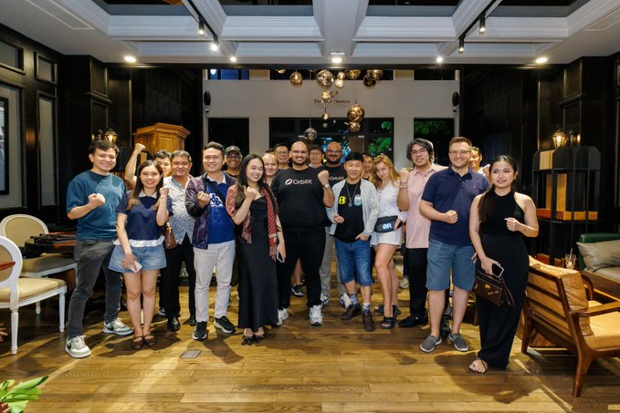 A group of people posing together in an elegant indoor setting, with one person wearing a shirt featuring the 'DogeX' logo.