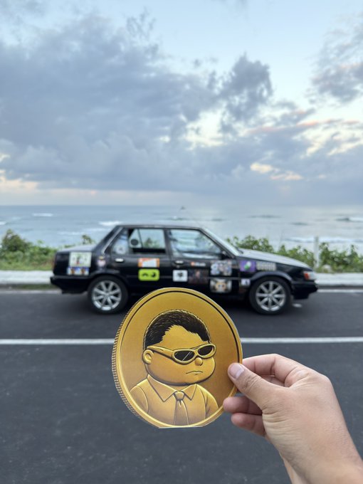 A hand holds a large gold coin featuring a cartoon character with sunglasses, with a sticker-covered black hatchback car parked by the ocean in the background.