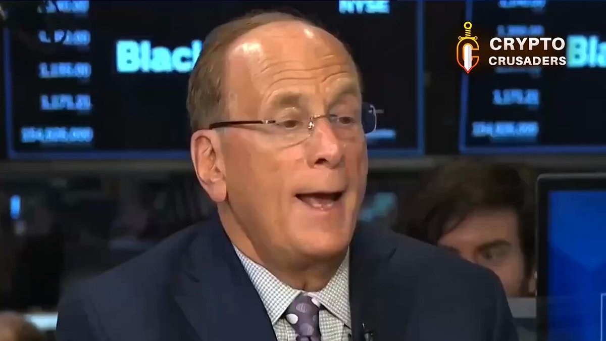 The image shows a man wearing glasses, talking into a microphone while sitting at a desk with computer monitors behind him. The background features multiple screens displaying stock market data, suggesting that he might be discussing financial matters or analyzing market trends related to Bitcoin.