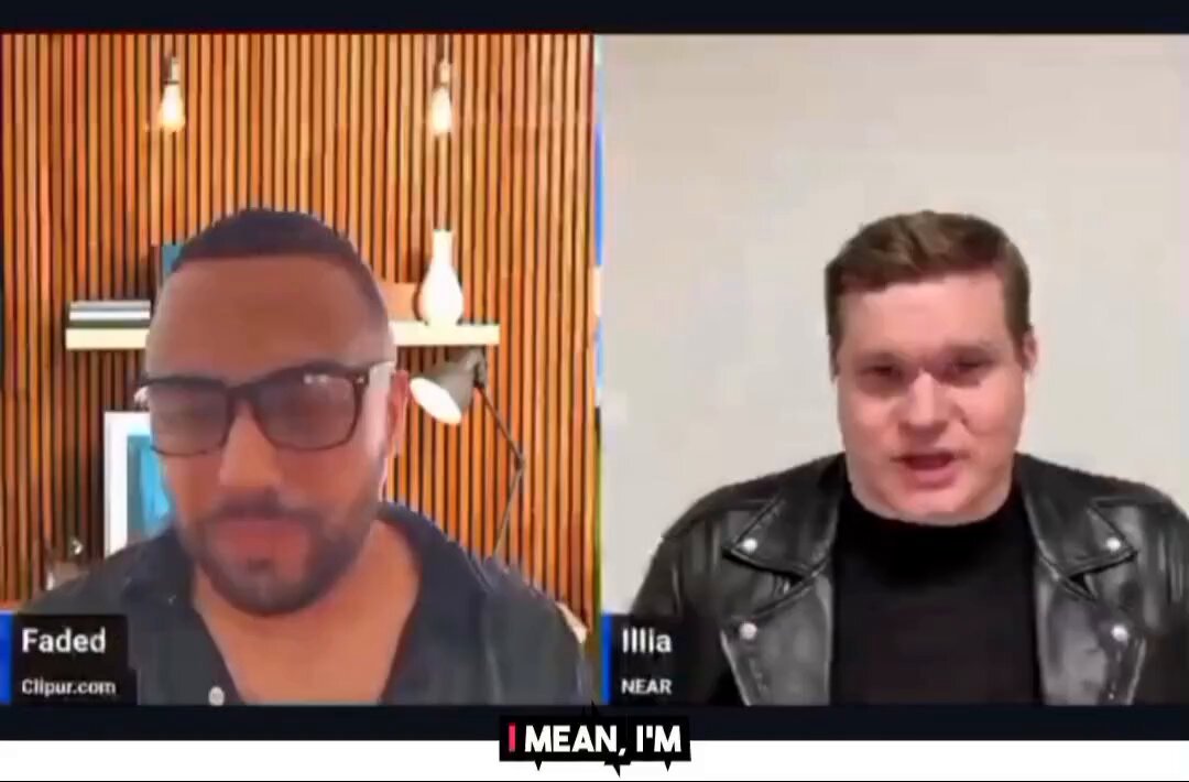 The image shows two men having a conversation over a video call. One man wears glasses and has a beard, while the other man is wearing a leather jacket. The background features a wooden wall with hanging lights that illuminate the scene. The caption of the image reads "I mean, I'm" which could be interpreted as an inside joke or a humorous statement made by one of the men during their conversation.