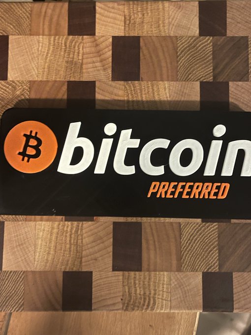 A black "Bitcoin Preferred" bumper sticker featuring the orange Bitcoin logo rests on a checkered wooden surface, symbolizing the poster's commitment to their BTC stacking strategy as they set and surpass annual accumulation goals ahead of 2026.