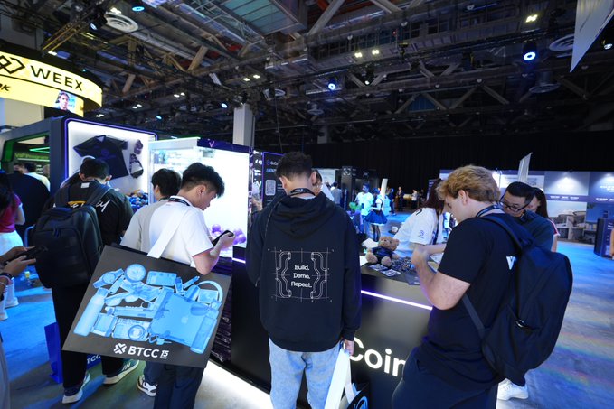 A scene from what appears to be a gaming or tech convention booth, with several people examining products on display, including someone carrying a BTCC-branded bag featuring a Gundam mecha design.
