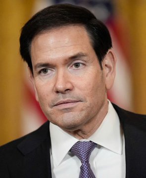 A close-up portrait of U.S. Secretary of State Marco Rubio wearing a dark suit and purple tie, with an American flag visible in the background.