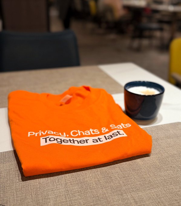 A folded orange shirt on a table reads 'Privacy, Chats & Sats — Together at last.' next to a dark mug of coffee.