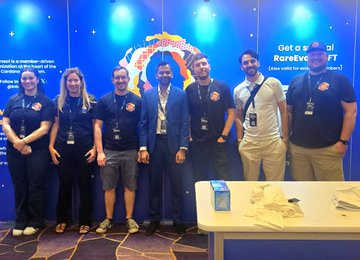 A group of seven people, including members of the Cardano community, pose together at the "RareEVO 2026" event, standing behind a table displaying promotional materials in front of a "Get a [Redacted] RareEvo NFT" backdrop. The image captures the collaborative spirit highlighted in the post, showcasing the networking and community connections that drive the blockchain ecosystem forward.