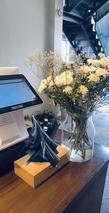 The image features a wooden desk with various items placed on top of it, including a vase filled with flowers and a computer monitor or cash register. The flowers are white and appear to be dried, adding an artistic touch to the scene. A small sculpture is also present on the desk, further enhancing the visual appeal of the space. The image seems to be related to a post about celebrating level #50 by buying Todd Batman b&w nft on veve or First DC by @veve_official, as indicated by the caption provided.