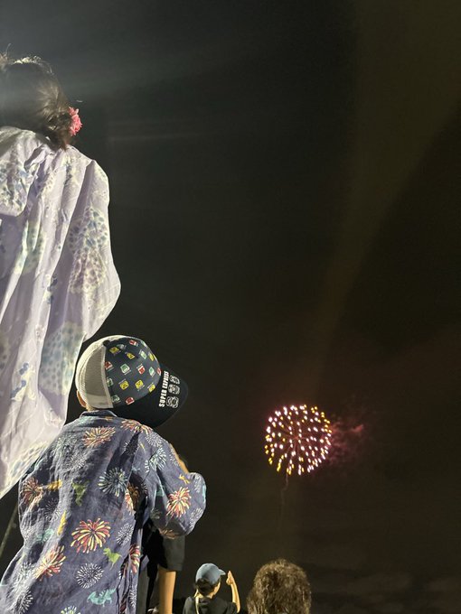 From behind, two people in traditional Japanese yukata stand watching a bright, glowing firework explode in the dark night sky.