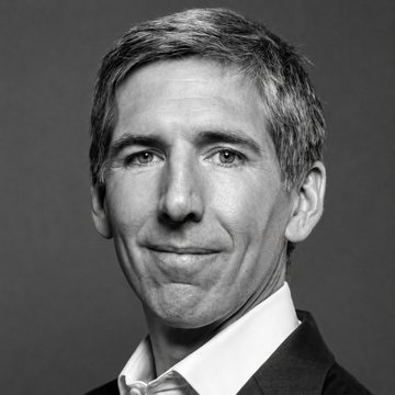 A black-and-white portrait photograph of a middle-aged man with short gray hair, wearing a white collared shirt, smiling slightly at the camera.