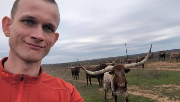 A man in an orange jacket takes a selfie in front of a herd of longhorn cattle in a rural, open landscape under a cloudy sky.