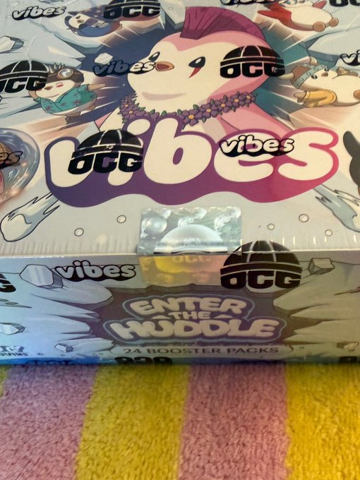 A sealed box of Pudgy Penguins Vibes TCG "Enter the Huddle" First Edition booster packs, containing 24 packs, sits on a pastel striped surface, matching the post about receiving this box and deciding whether to open it.