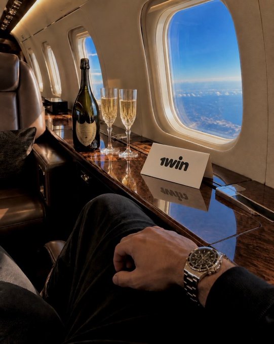 Luxury private jet interior with champagne bottle, two flutes, and a '1Win' card on a wooden console, alongside a man's hand wearing a watch.