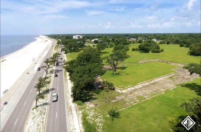 An aerial view of a coastal landscape showing a paved highway running alongside a white sand beach, with cars driving on the road, bordered by green marshlands, trees, and scattered houses under a blue sky.