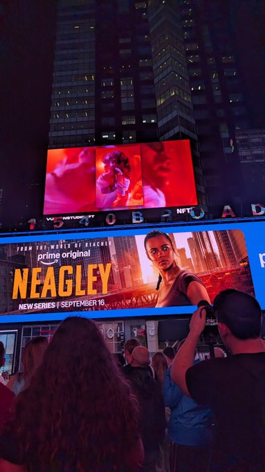 A nighttime crowd watches large digital billboards in a city, including a prominent ad for the Prime Original series 'NEAGLEY' premiering September 16, while another screen above shows a red-tinted image.