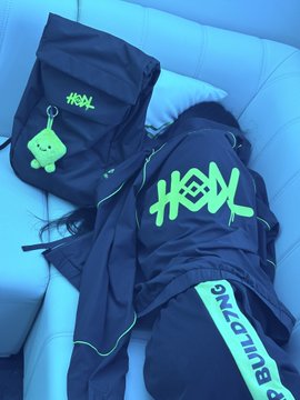 Black streetwear clothing and accessories featuring bright green "HEXL" branding, including a cap with a green alien keychain and a hoodie with text reading "B U M C M B K."