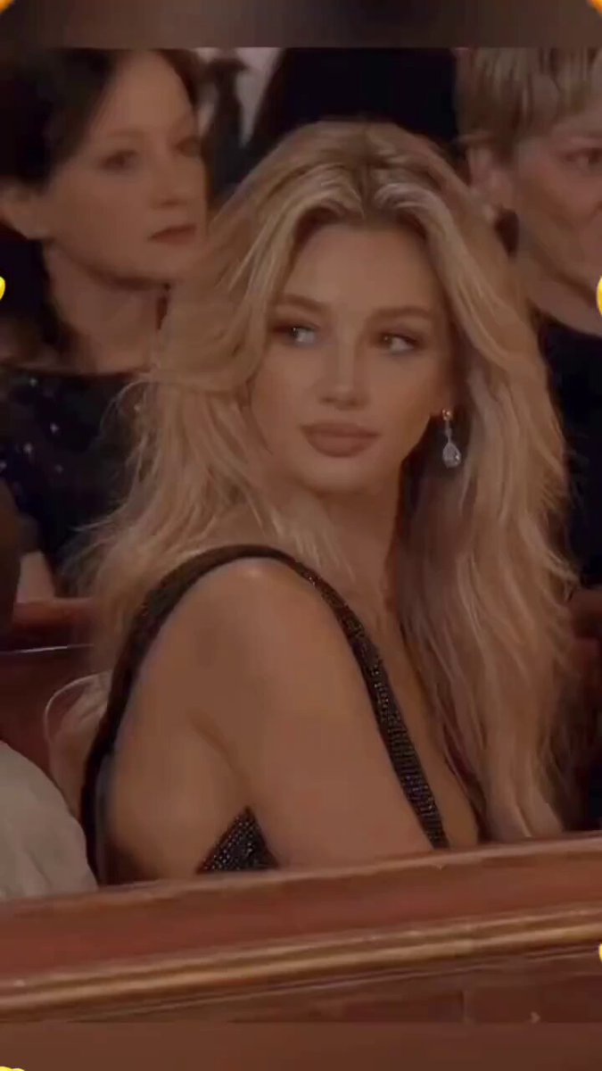 The image features a woman with blonde hair wearing a black dress sitting in front of an audience at a concert or event. She appears to be looking off into the distance, possibly lost in thought or daydreaming. The background shows other people seated around her, indicating that she is not alone in this setting. There are also some chairs visible in the scene, suggesting that it might be a venue for an event or gathering.