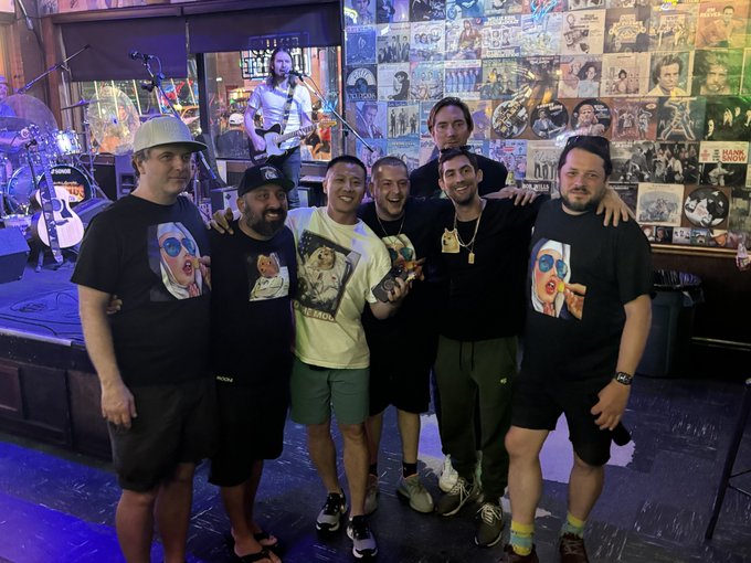 A group of seven men posing together in a bar/venue with walls covered in music memorabilia and posters, wearing casual clothing including graphic t-shirts and caps.