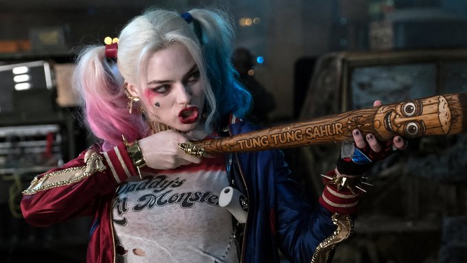 The image features a woman with purple hair wearing a colorful t-shirt holding a wooden baseball bat, seemingly ready for action. The scene also includes text related to a popular movie franchise or series called "TripleT", which refers to the actress playing Summer Sahur in the film "The Despicable Me". This indicates that her character is likely preparing for an upcoming role or event within the franchise.