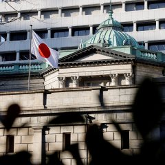 The Bank of Japan building with its distinctive green dome and the Japanese national flag flying in front, partially obscured by a dark silhouette in the foreground.