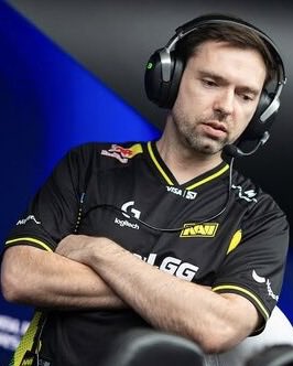 A photo of a bearded esports player wearing a headset and a black and yellow NAVI jersey with logos including Logitech and Visa.
