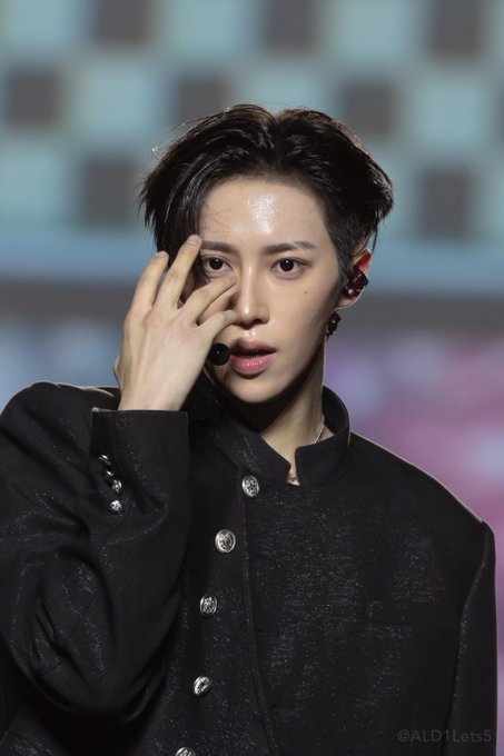 A close-up photo of a performer with short dark hair wearing a black button-up jacket and in-ear monitor, captioned '@ALDI_Lets5'.