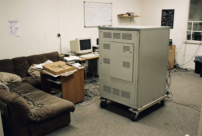 An old office or dorm room containing a large beige server cabinet on wheels, a desk with a vintage CRT monitor, and a worn brown sectional sofa.