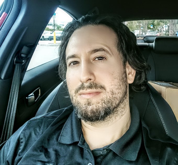 A close-up selfie of a man with dark wavy hair and a beard sitting in the driver's seat of a car, wearing a dark polo shirt, with a cardboard box visible on the passenger seat.
