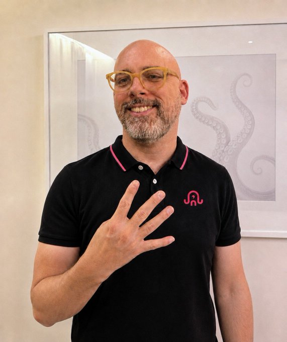 A bald man with a gray beard and yellow-framed glasses smiles and holds up four fingers while wearing a black polo shirt with a pink logo and pink collar trim.