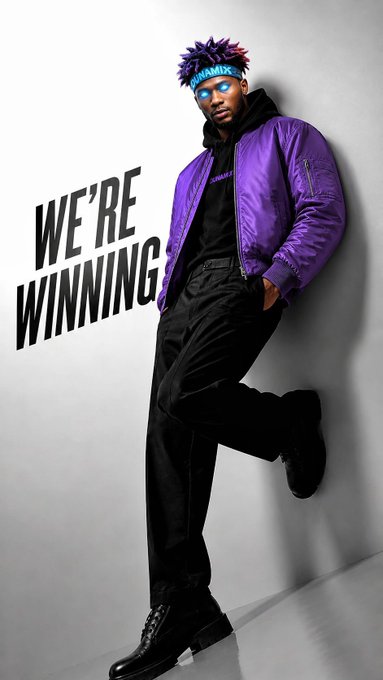 A man with a purple spiky hairstyle wearing a shiny purple bomber jacket and black pants, leaning against a wall with the text 'WE'RE WINNING' beside him.