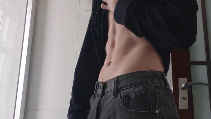 A person wearing a black cropped hoodie and dark gray jeans, showing their midriff, standing indoors near a door.