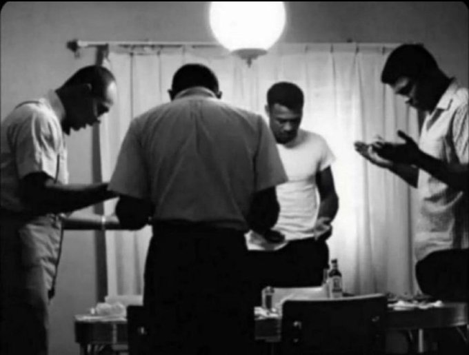 A vintage black-and-white photograph of four men gathered around a table, appearing to be playing cards or examining something in their hands.