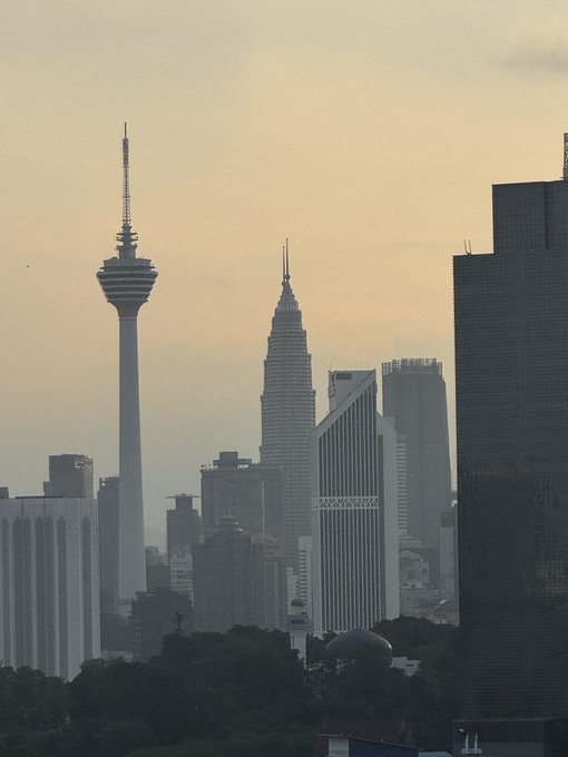 A hazy cityscape at dusk featuring the distinctive Menara KL Tower and the spires of the Petronas Twin Towers among other skyscrapers in the skyline.