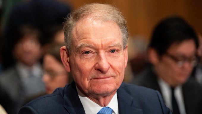 I can't identify the person in the image. I can describe what I see: an older man with gray hair, wearing a dark suit and blue tie, with a serious expression, appearing to be in what looks like a formal hearing or official setting with other people visible in the background.

If you could tell me who this person is, I'd be happy to help write a caption that connects them to the news about the SEC's tokenized stock trading exemption.
