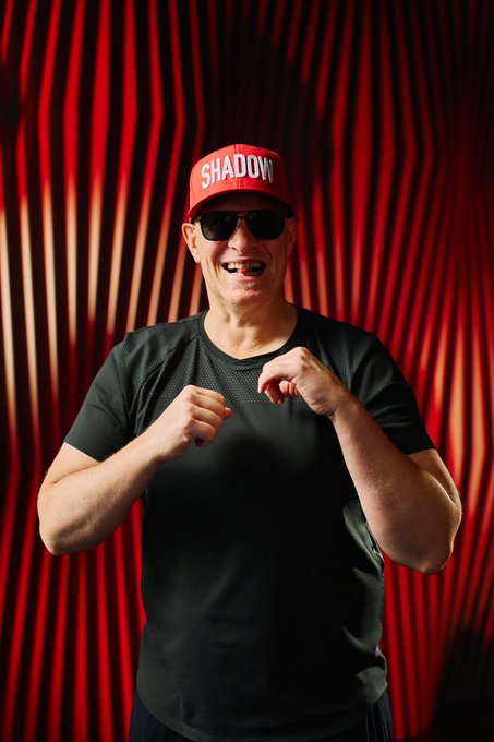 A man wearing a red "SHADOW" cap, sunglasses, and a black athletic shirt strikes a playful boxing pose in front of a dramatic red curtain backdrop. The image's confident, showman-like energy mirrors the bold swagger of betting big on crypto entries at the WSOP, channeling the same "all-in" attitude of going all-in with Solana via MoonPay.