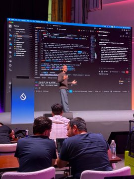 A speaker addresses an audience at the SuiBasecamp event, presenting code on a large screen behind him, as attendees listen attentively while seated at tables with water bottles. The image captures the technical session described in the post, where D. Lougaris discusses agent memory architecture and leads a workshop on building agents with persistent memory capabilities.