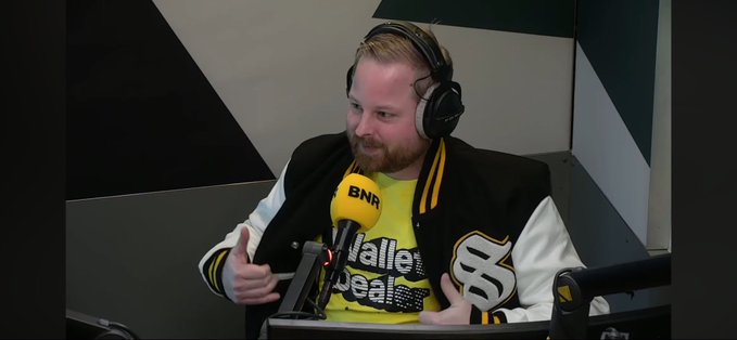 A bearded man wearing a "BNR" headset microphone and a black and yellow varsity-style jacket appears to be hosting a radio or podcast segment, giving a thumbs-up while speaking. The "BNR" logo on the mic likely connects to the post's mention of "Solflare," suggesting this is a broadcast promoting the Solflare crypto wallet platform.