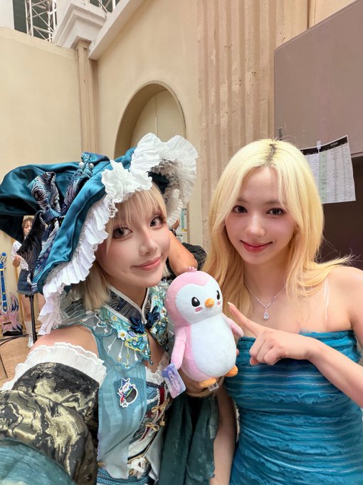 Two women pose together indoors; one wears a frilly blue and white maid-style outfit, while the other has blonde hair and a blue dress, holding a pink plush penguin between them.