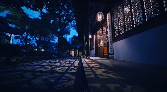 A nighttime photograph of a traditional Chinese garden corridor with intricately patterned lattice windows, glowing red lanterns, and long shadows cast across the stone pathway.