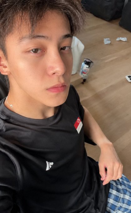 The image features a young man wearing a black shirt with a white logo, sitting down and looking at the camera. He has short hair and is dressed casually for the occasion. The background of the photo shows a wooden floor and a chair nearby. There are also some items scattered around him, including a bottle on the floor to his right and a cell phone resting on it.