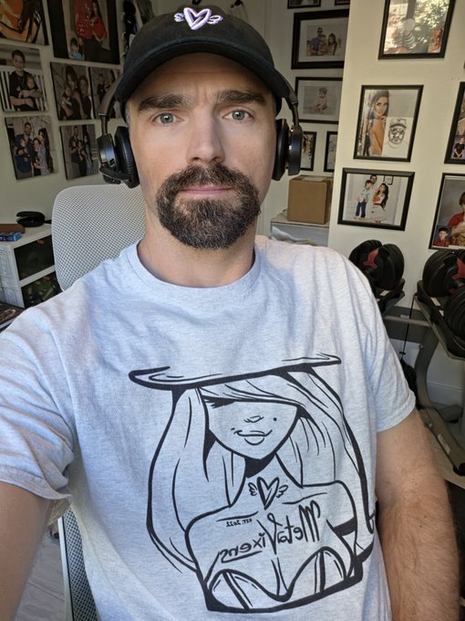 A bearded man wearing a black cap and headphones sits in a room, with a gray t-shirt featuring a stylized illustration and the text 'all dressed' worn by the figure.