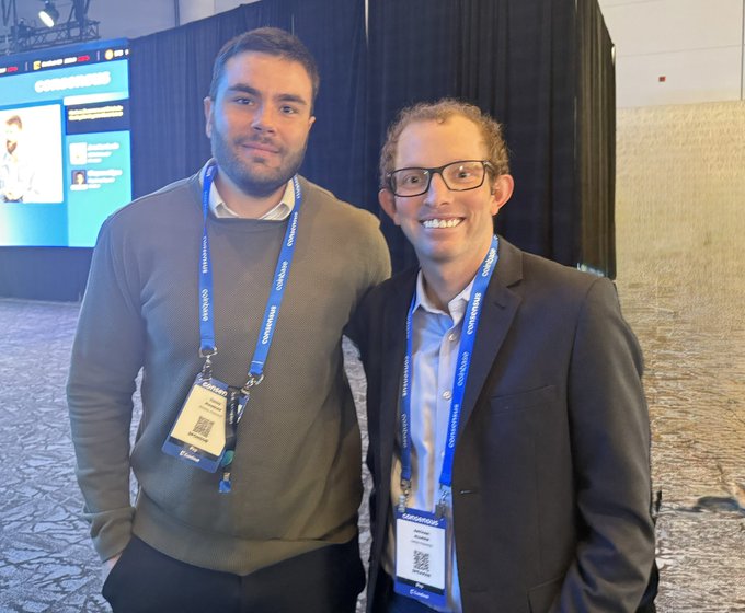 The image shows two men standing next to each other with their name tags attached to their lanyards. They are both smiling at the camera, creating an engaging atmosphere for the photo. The background of the image features a large screen displaying information about tokenization and digital assets, which may be related to the event they are attending or the post they have created.