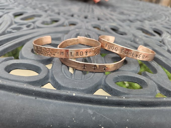 Three copper-colored cuff bracelets with hand-stamped text reading 'MILADY' and 'FOREVER' alongside decorative cross and diamond marks, resting on a black ornamental metal table.